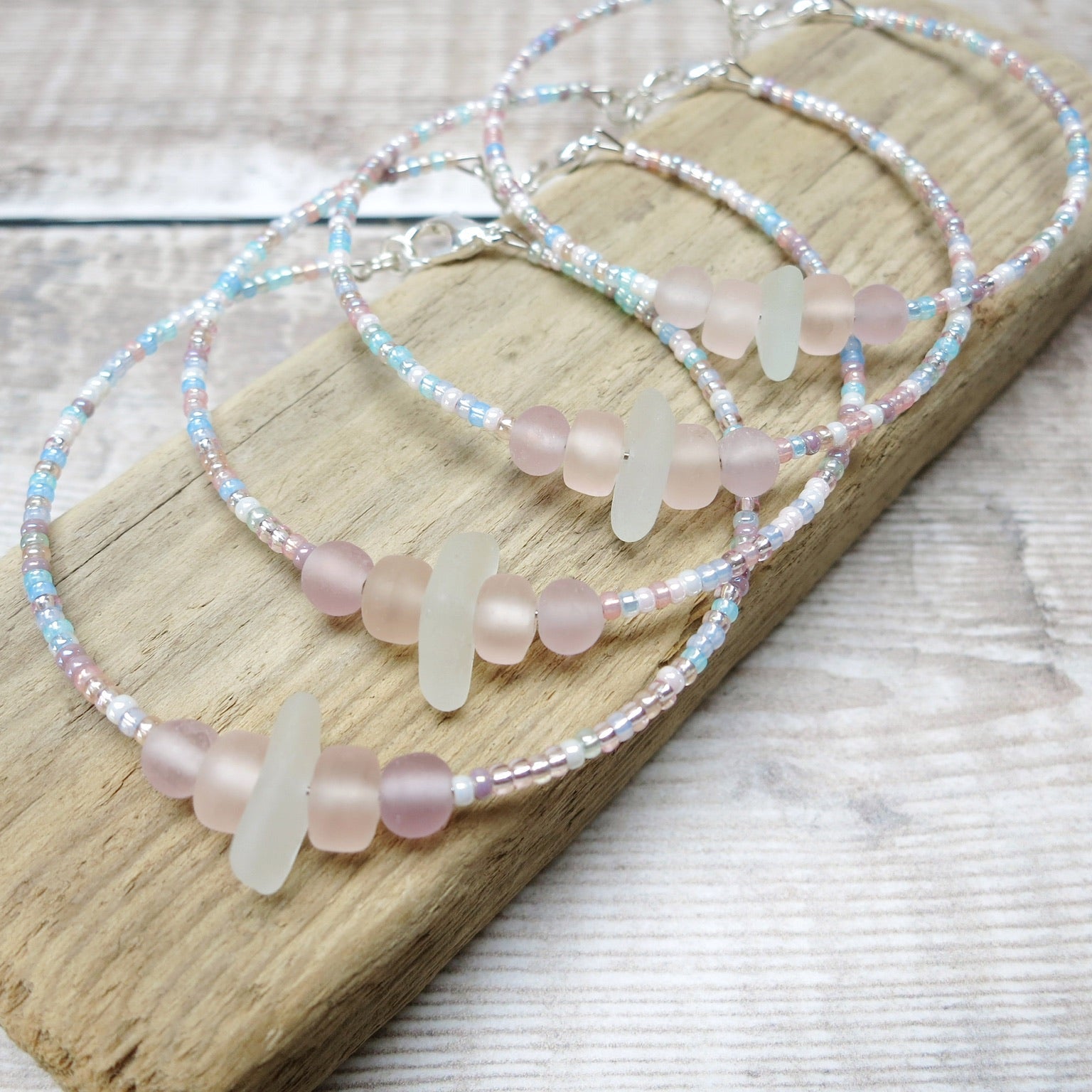 Four bracelets with white sea glass and pastel beads laid on a purple pebble against a wooden background
