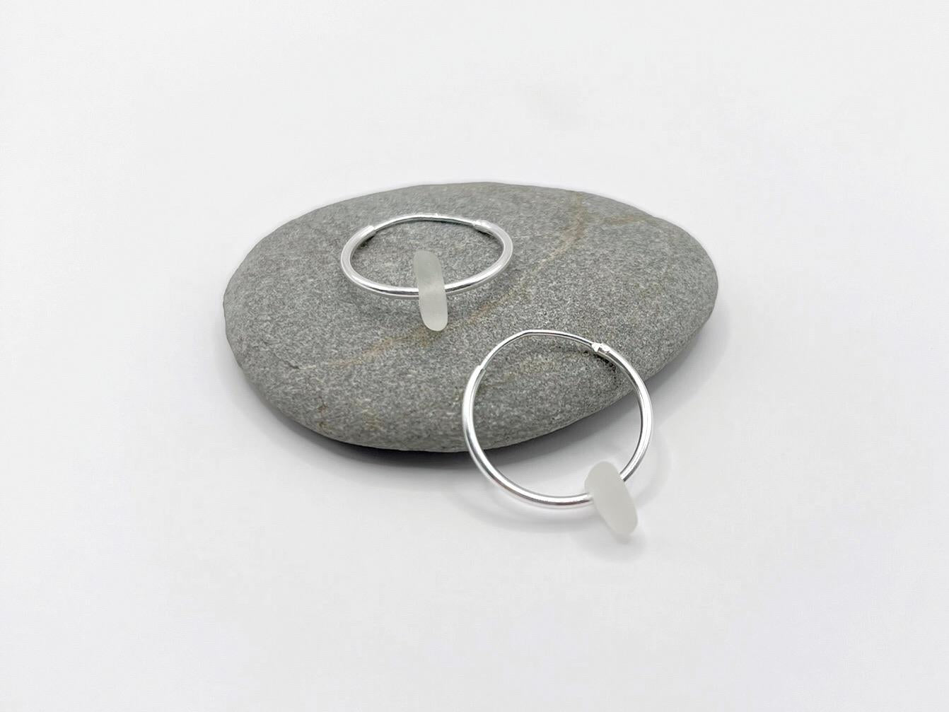 Silver hoop earrings with white sea glass laid on a grey pebble against a white background