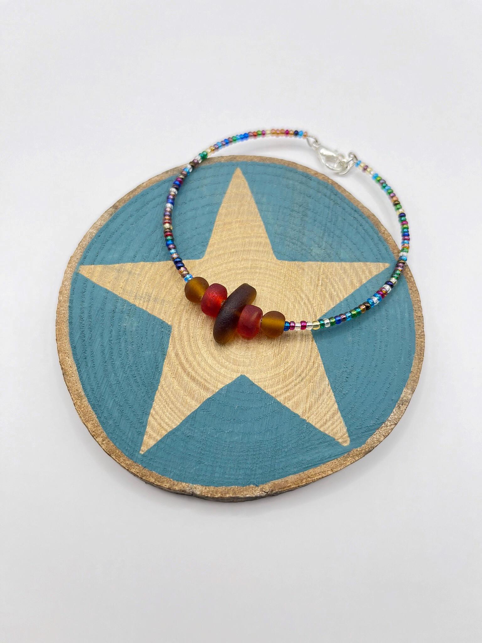 Rainbow bead bracelet with sea glass and red and brown frosted glass beads laid on a slice of wood with a star print