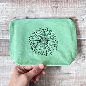 A mint green fabric pouch with a black lino print of a daisy design on the front and a zipper closure. Held in a hand against a wooden background.