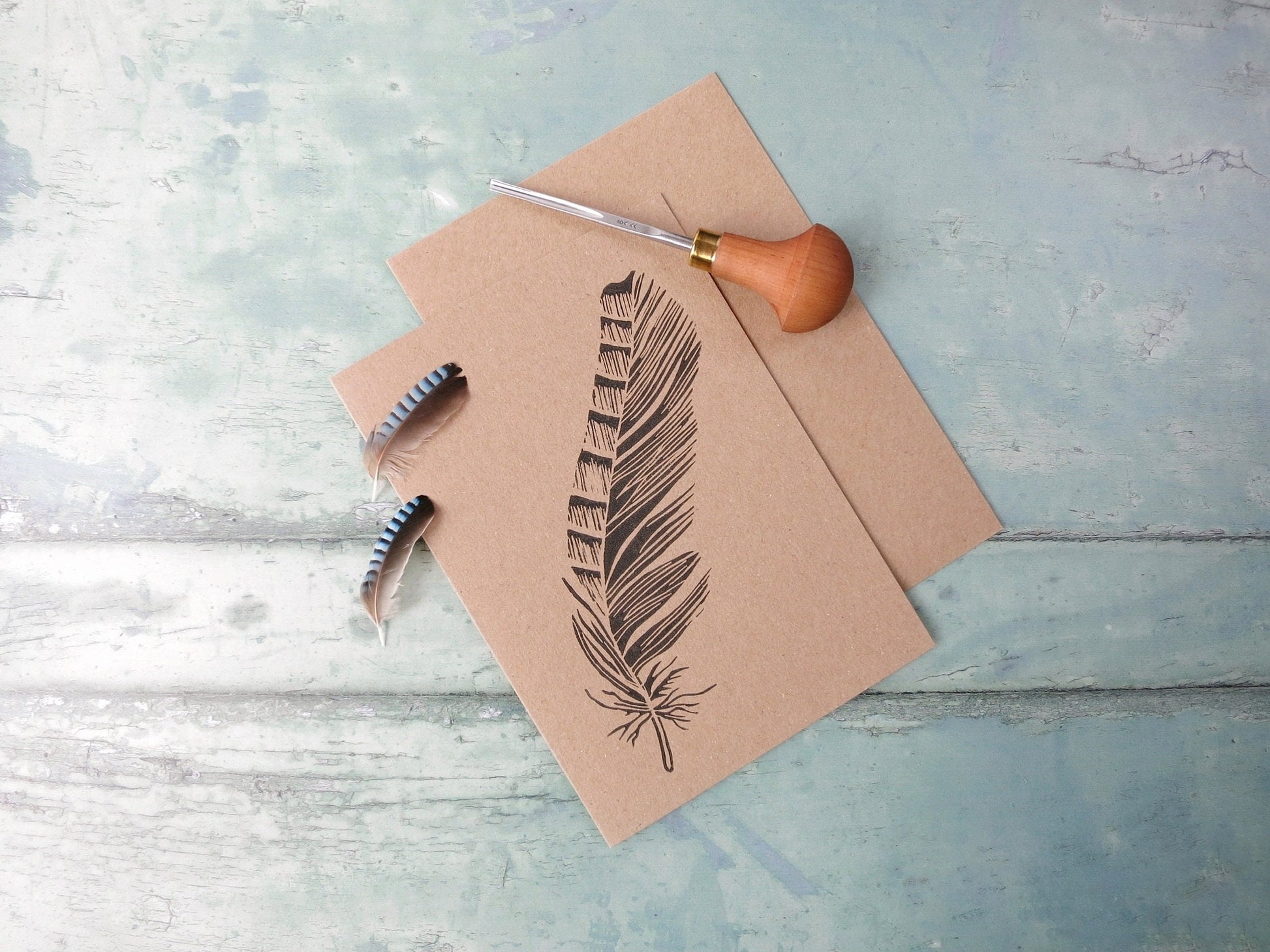 Kraft paper card with original lino print feather design. Included in the photo is the lino cutting tool used and two jay feathers