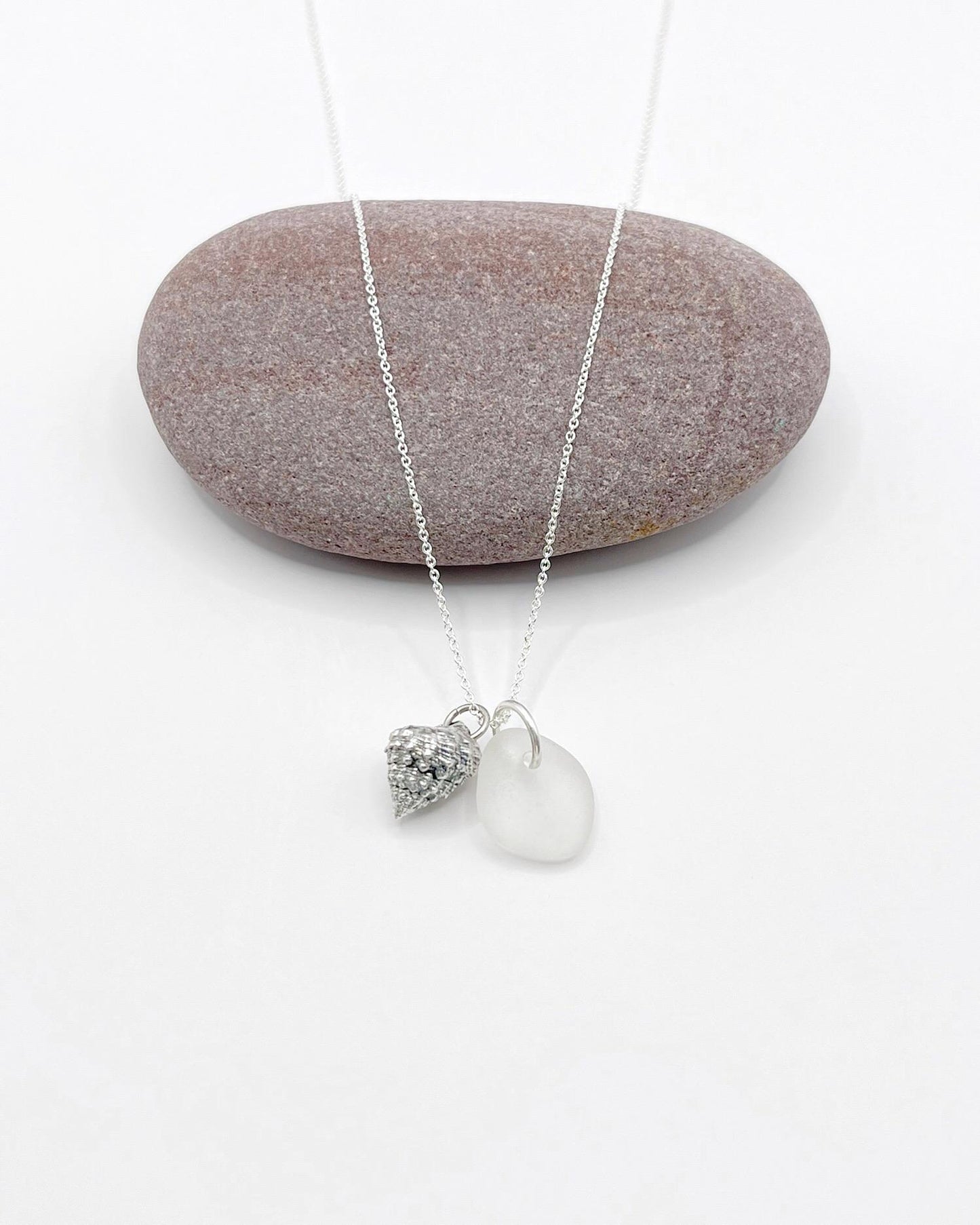 Silver necklace with White Sea glass and silver shell pendant laid in front of a purple pebble against a white background