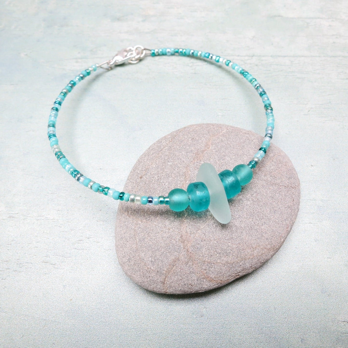 Bracelet with pale sea foam sea glass and sea green and turquoise glass beads laid on a grey pebble against a pale blue background