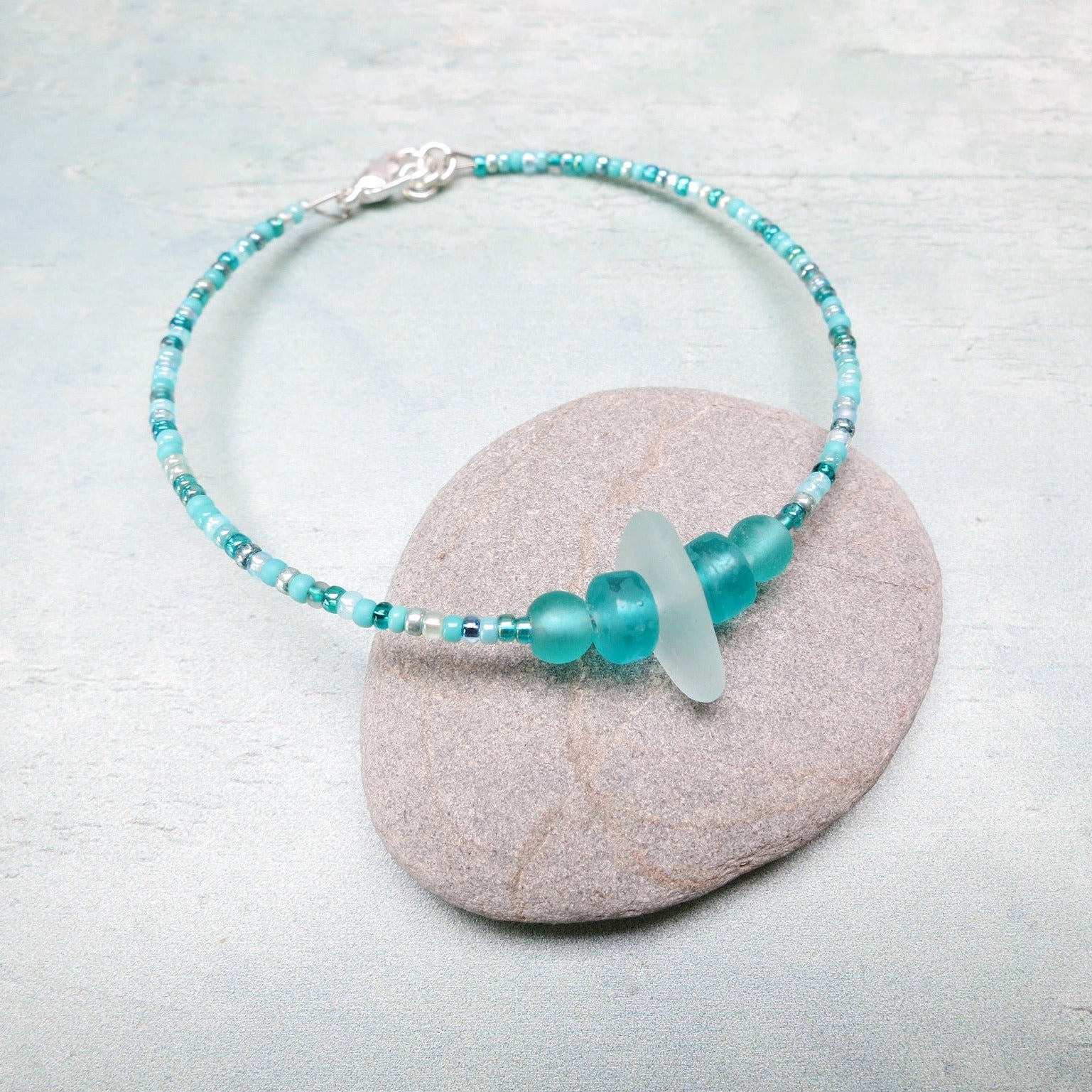 Bracelet with pale sea foam sea glass and sea green and turquoise glass beads laid on a grey pebble against a pale blue background