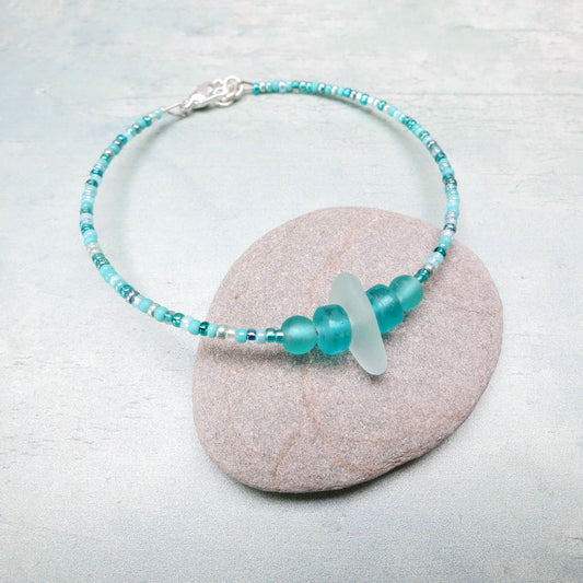 Bracelet with pale sea foam sea glass and sea green and turquoise glass beads laid on a grey pebble against a pale blue background