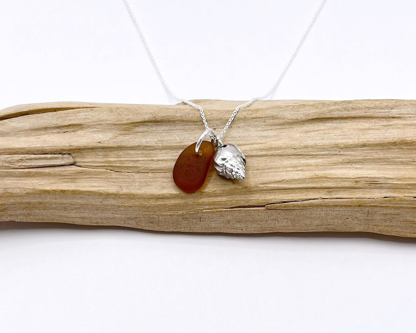 Necklace with burnt orange sea glass and silver shell pendant laid on driftwood against a white background