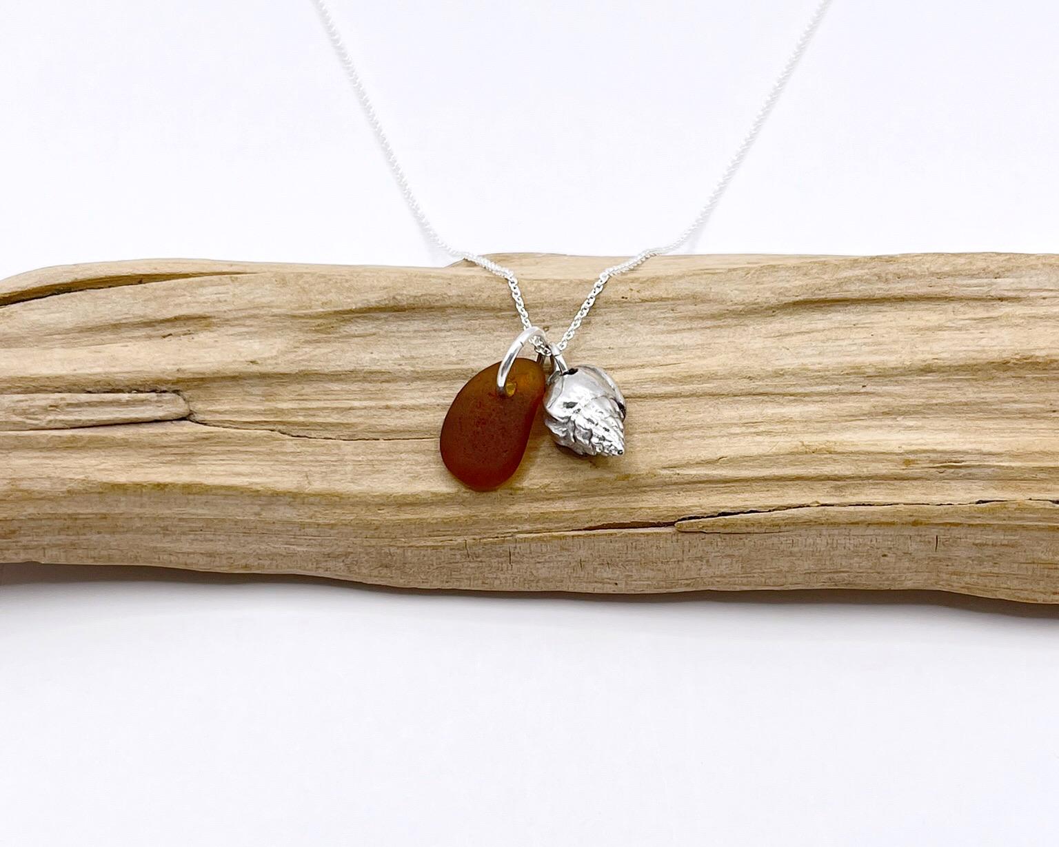 Necklace with burnt orange sea glass and silver shell pendant laid on driftwood against a white background