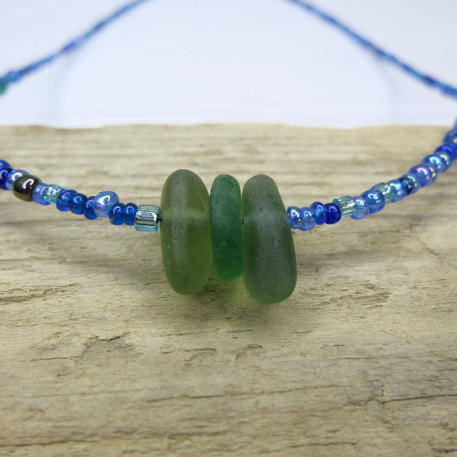 Close up of blue seed bead necklace with green sea glass laid on driftwood.