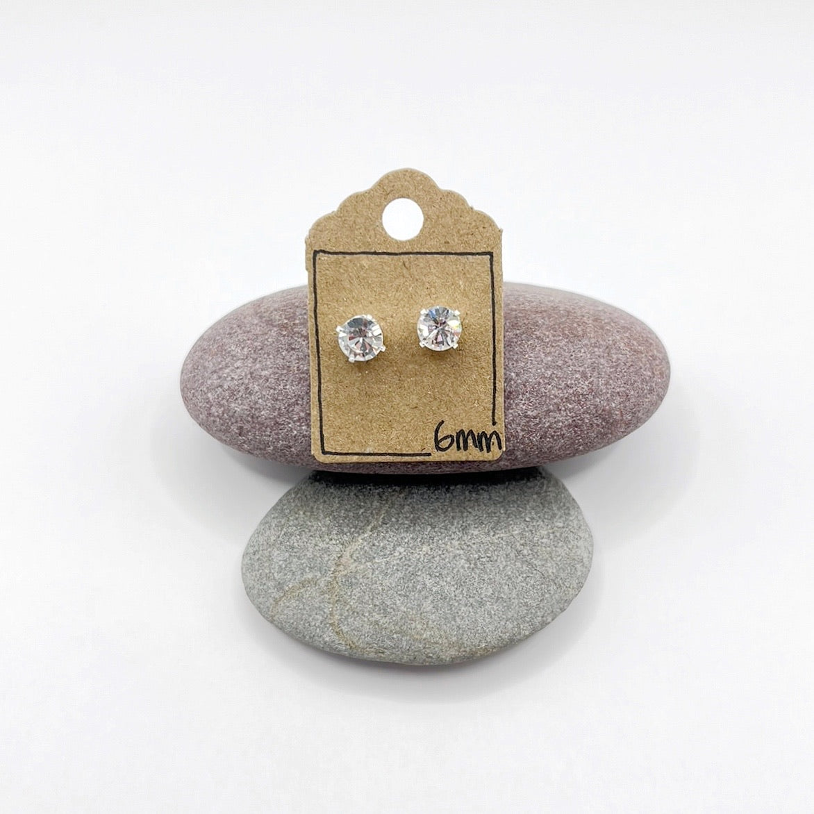 Silver stud earrings with clear cut glass on a cardboard backing card with '6mm' text, placed on two stones.