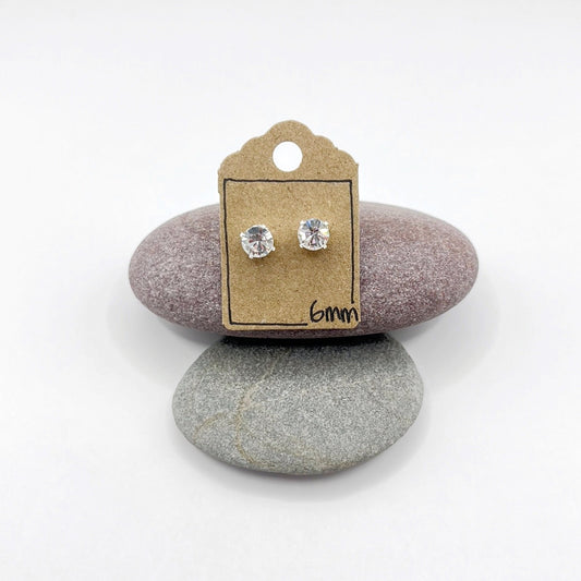 Silver stud earrings with clear cut glass on a cardboard backing card with '6mm' text, placed on two stones.