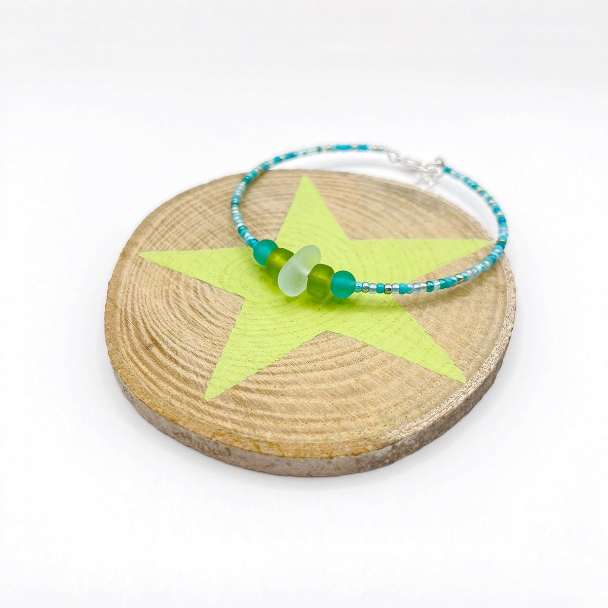 A turquoise beaded bracelet with lime and sea green frosted glass beads and a piece of sea foam coloured Cornish sea glass, finished with a silver plated lobster clasp and laid on a piece of wood with a pale lime star design.