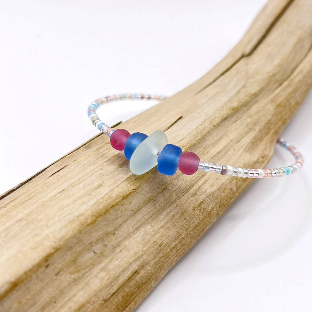 A pastel beaded bracelet with pink and blue frosted glass beads and a piece of sea foam coloured Cornish sea glass, finished with a silver plated lobster clasp and laid on a piece of driftwood.