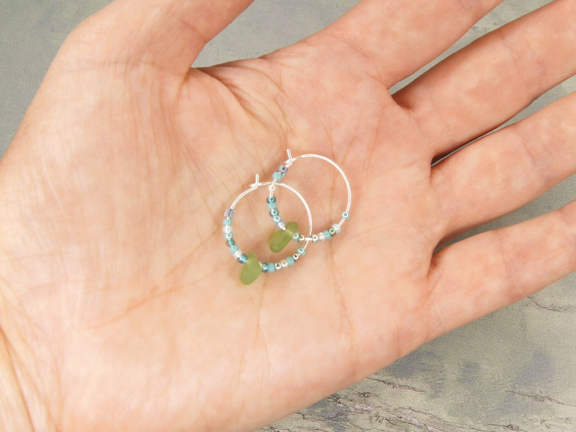 Two small turquoise seed bead hoop earrings with green sea glass held in a hand against a neutral background