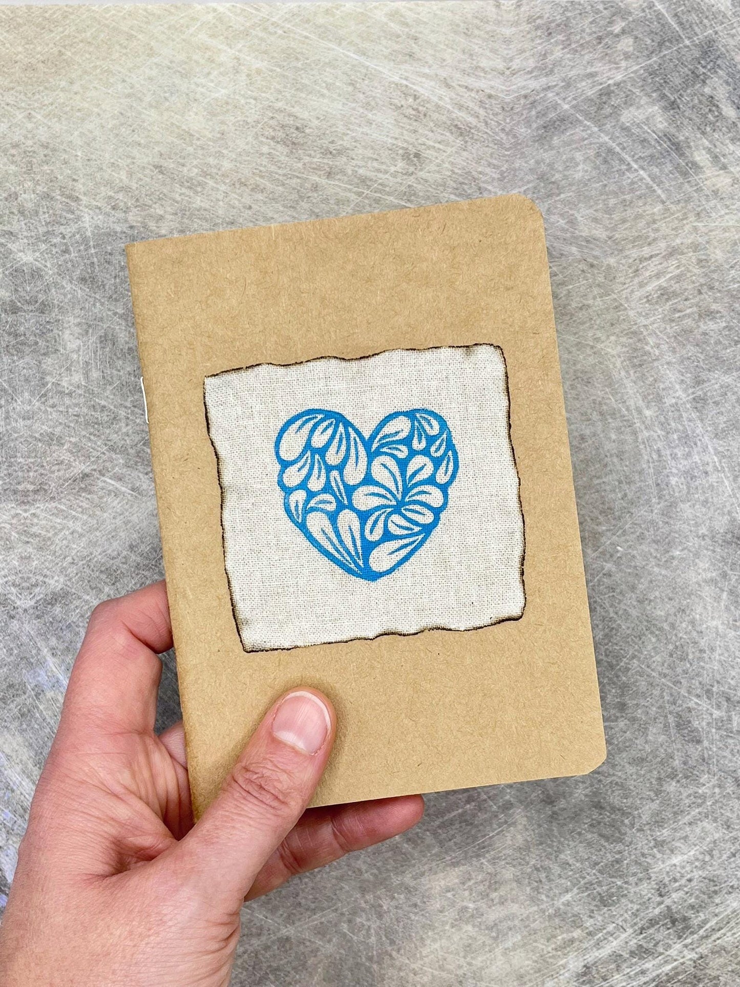 Hand holding a small brown notebook with a floral heart design on grey surface