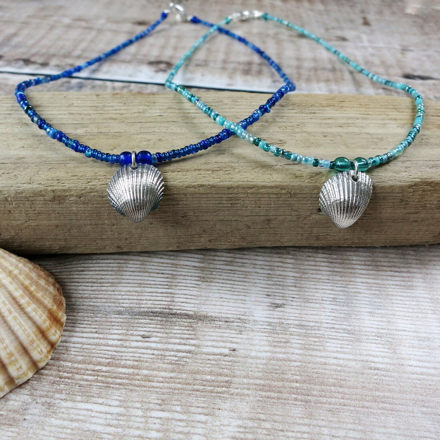 Two seed bead necklaces with silver coated shell pendants laid on driftwood against a textured surface