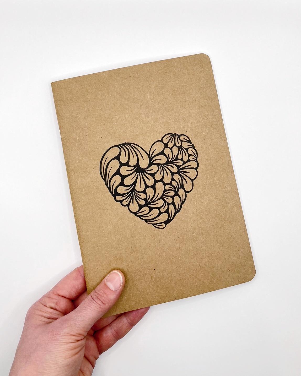 A5 Kraft sketchbook with original lino printed heart held in a hand against a white background