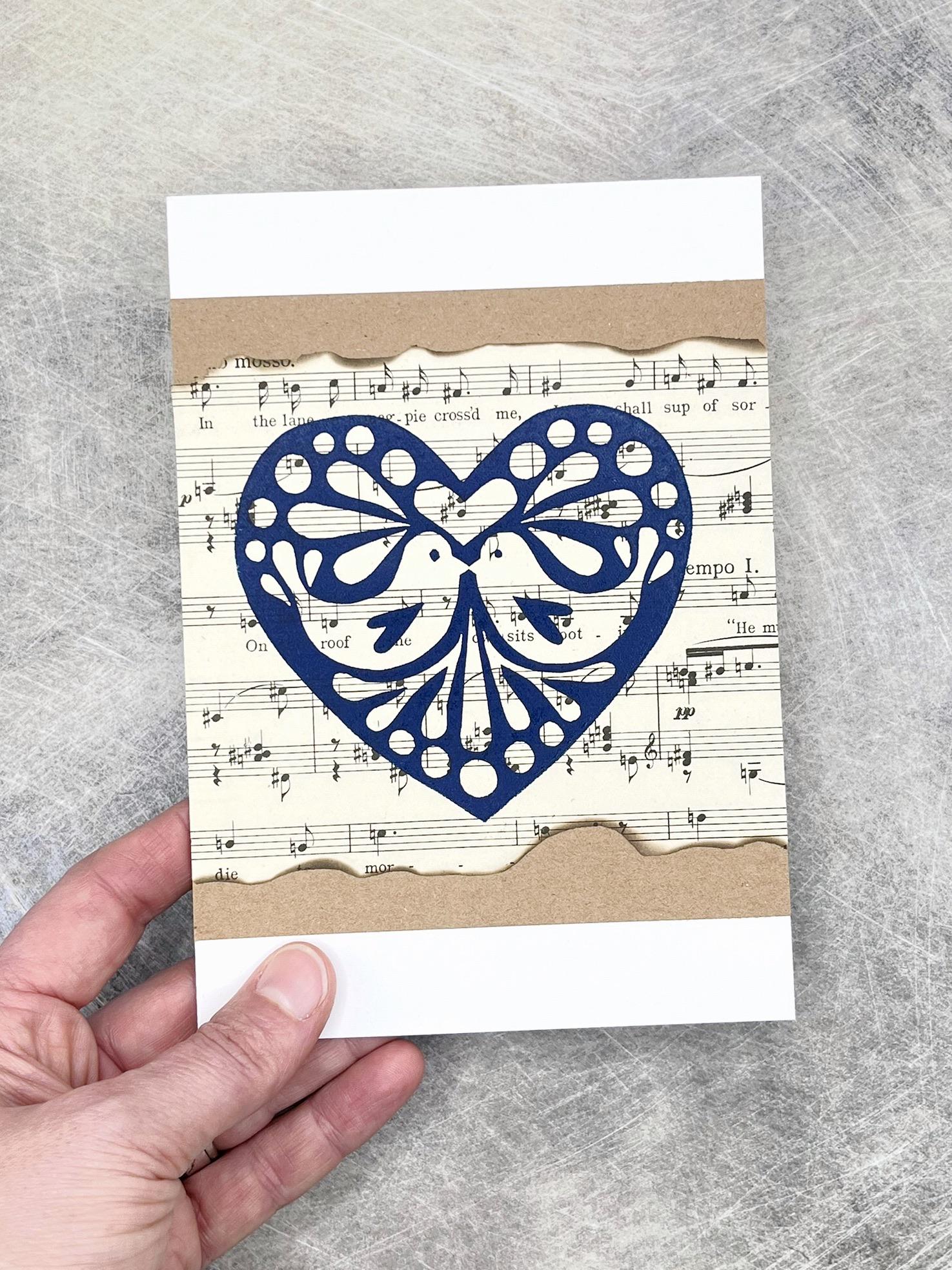 Card with blue heart design on sheet music held by a hand against a grey textured background