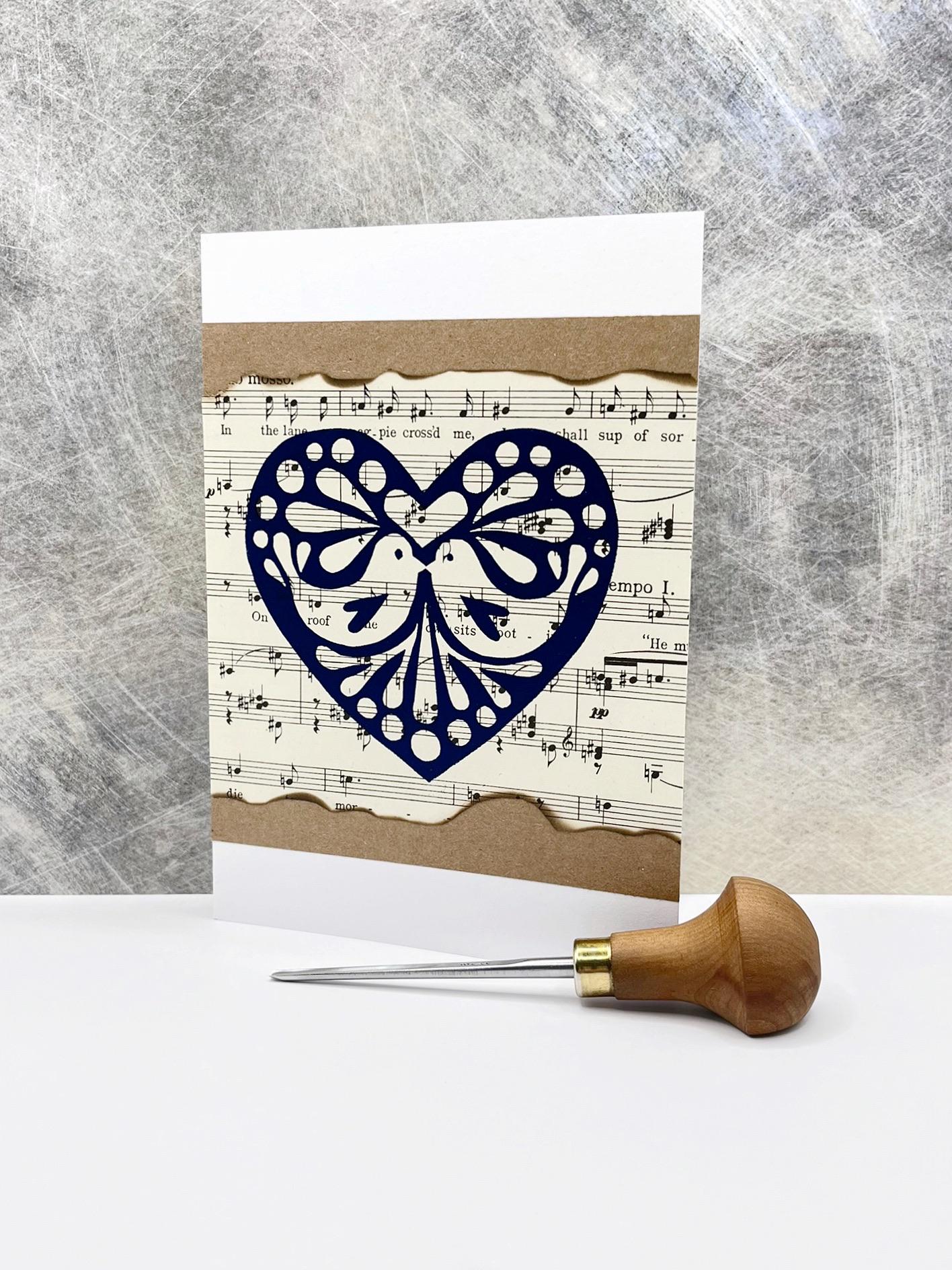 Card with original lino printed blue heart on sheet music. Photo includes lino cutting tool stood against a textured background.