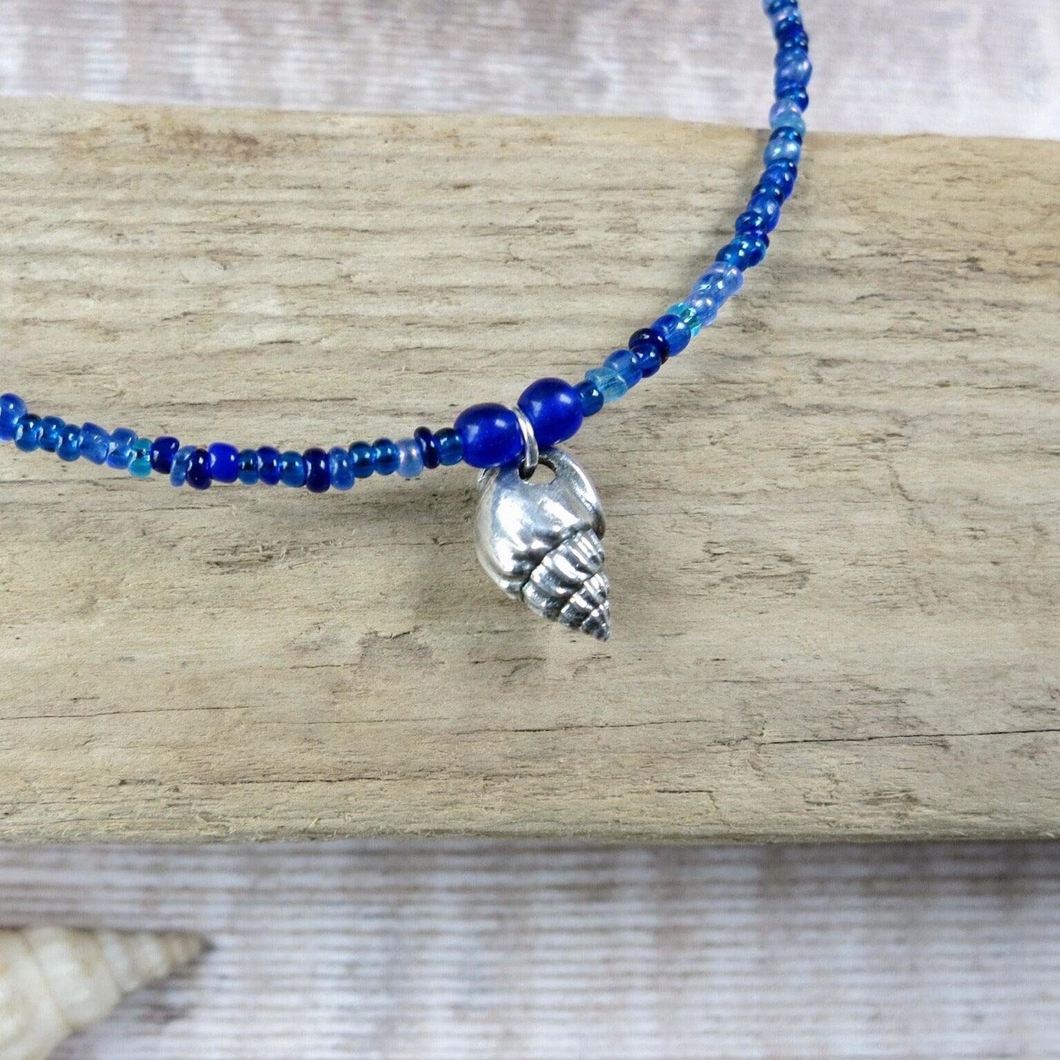 Blue seed bead necklace with a silver shell pendant laid on driftwood against a textured surface