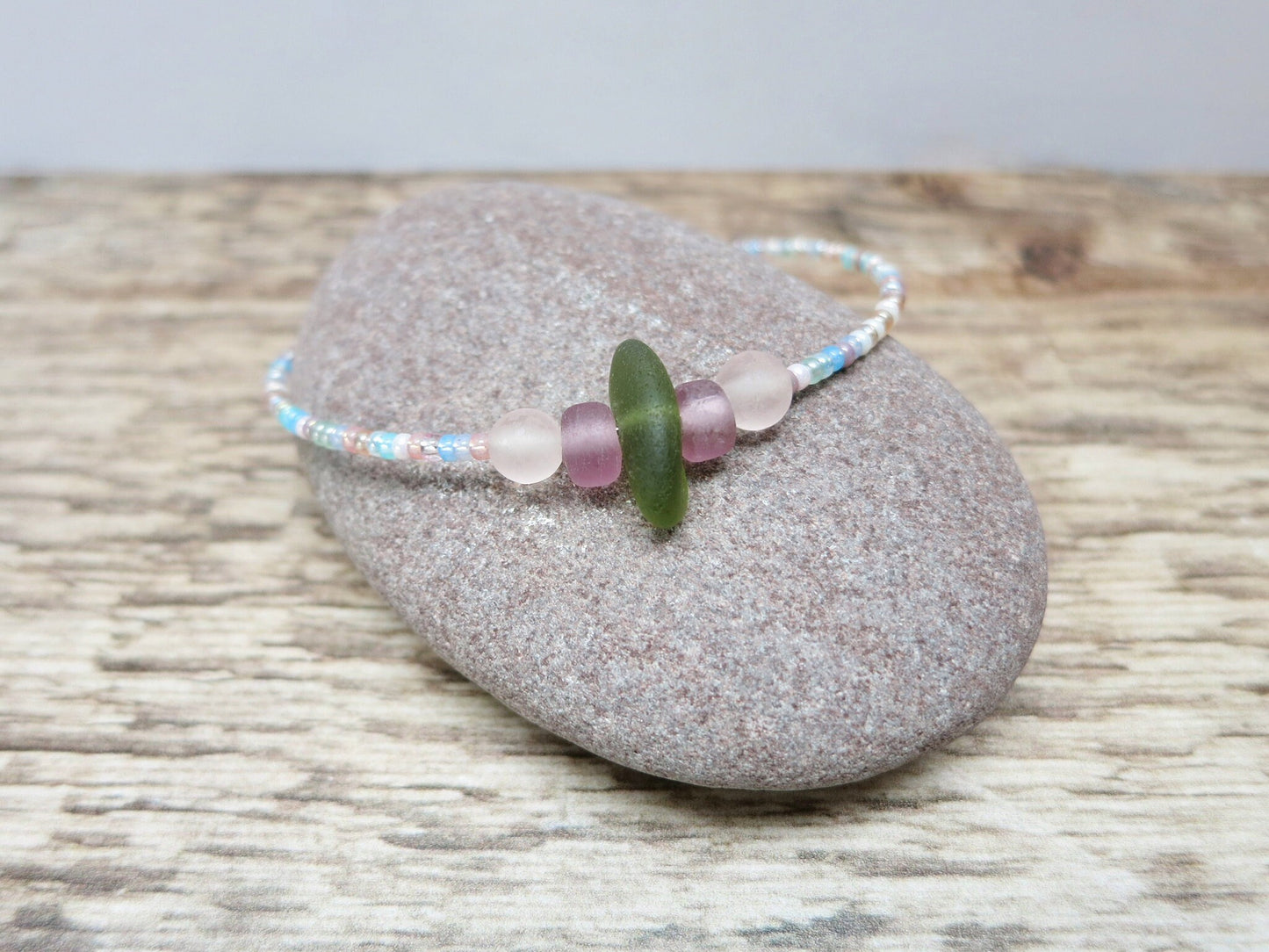 Pastel seed beaded bracelet with olive green sea glass laid on a pebble on a wooden surface