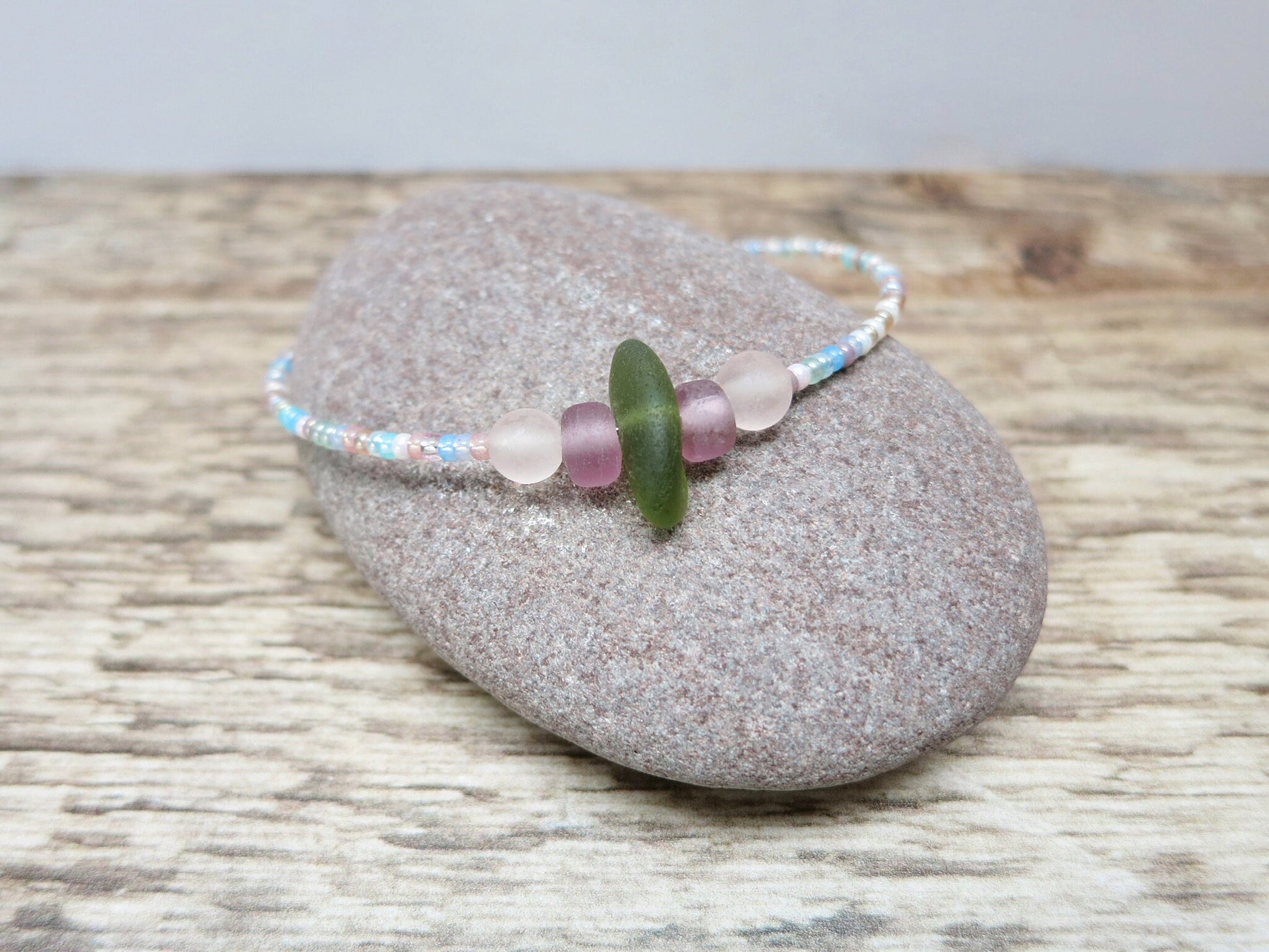 Pastel seed beaded bracelet with olive green sea glass laid on a pebble on a wooden surface