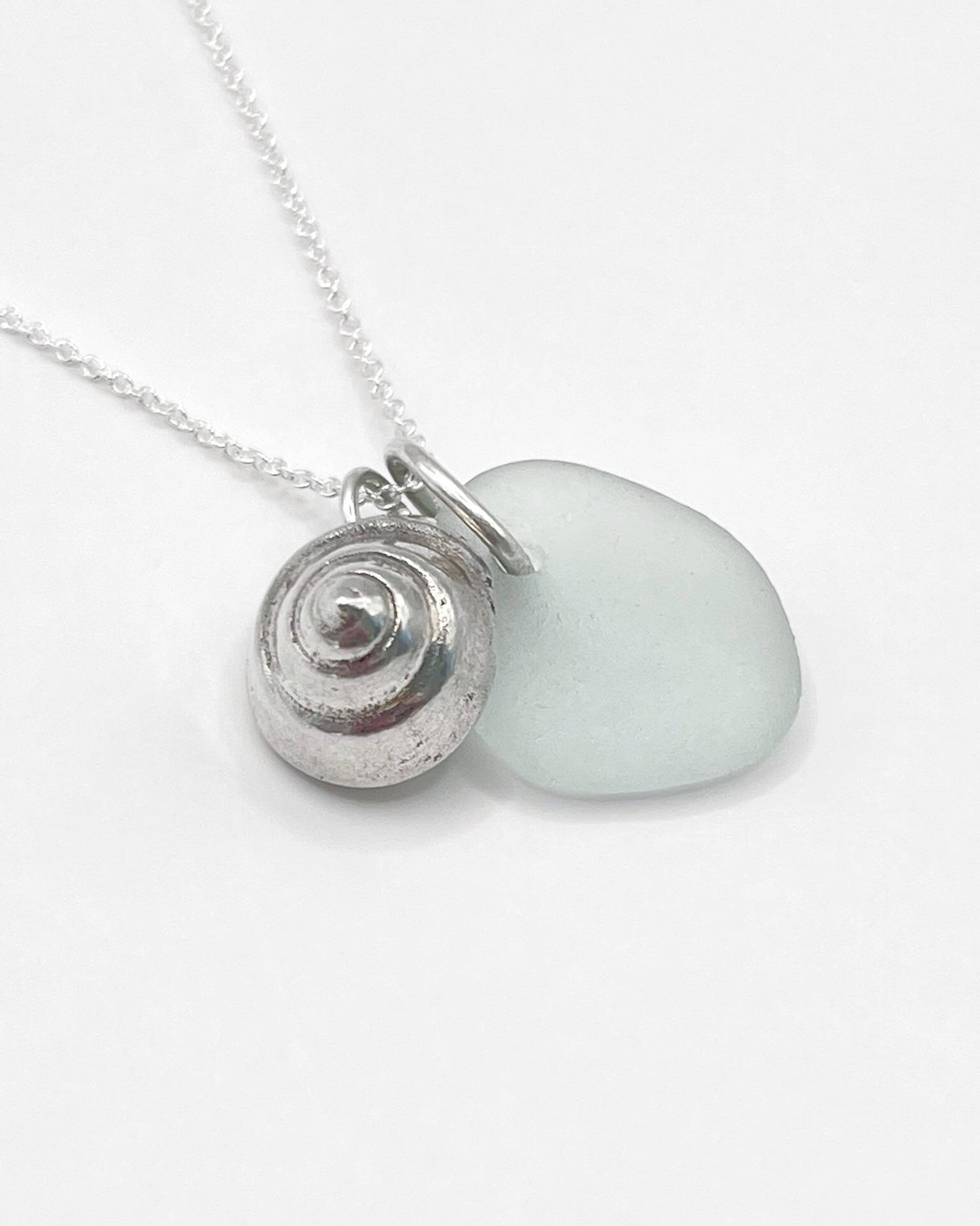Close up of necklace with a silver shell and seaglass pendant laid against a white background