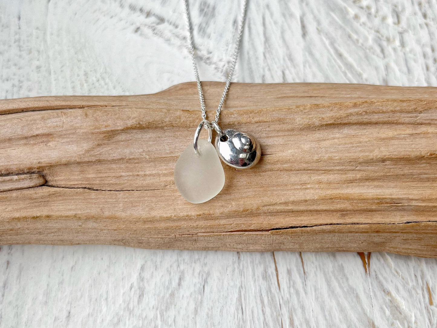 Necklace with a silver shell and white sea glass pendant laid on driftwood against a textured surface