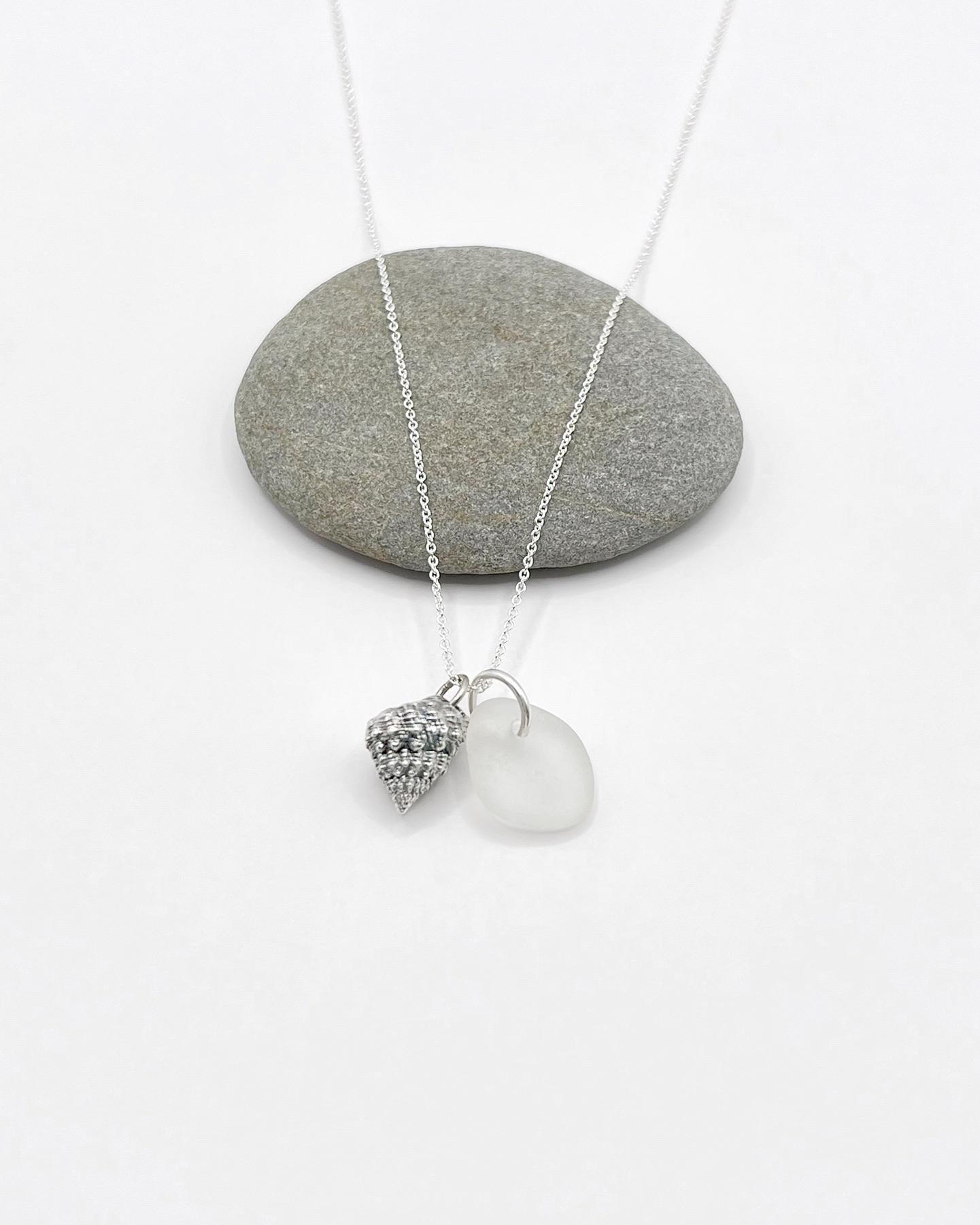 Silver necklace with White Sea glass and silver shell pendant laid in front of a grey pebble against a white background
