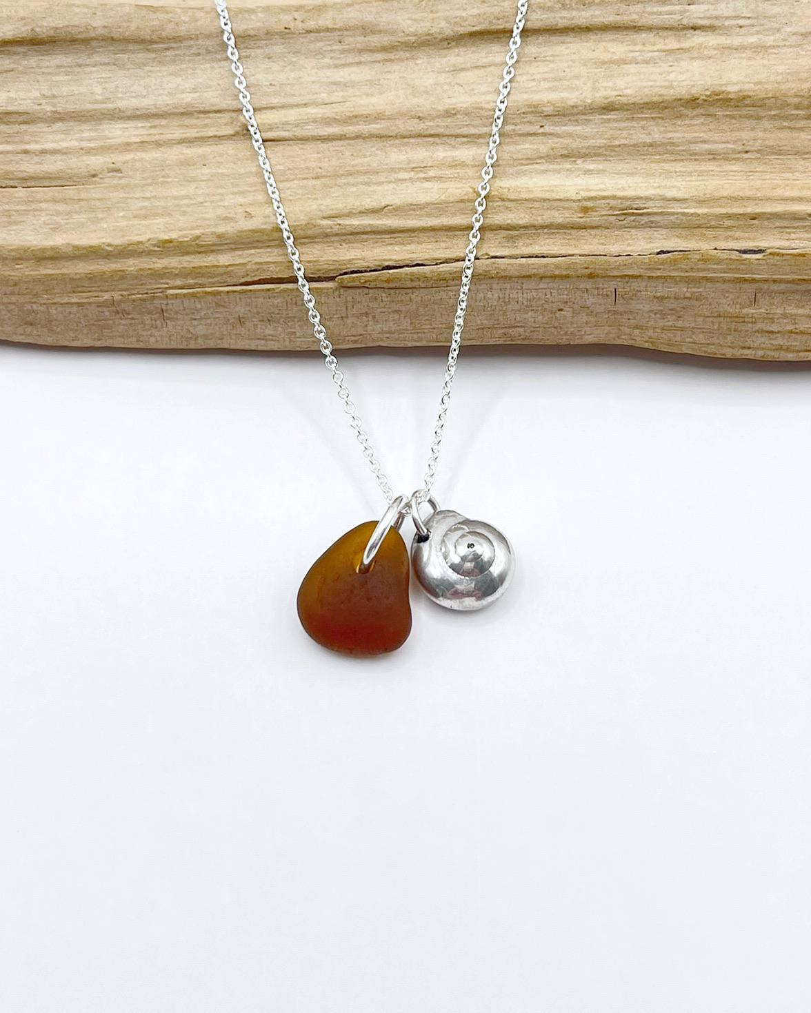 Necklace with burnt orange sea glass and a silver shell pendant, laid in front of driftwood on a white surface