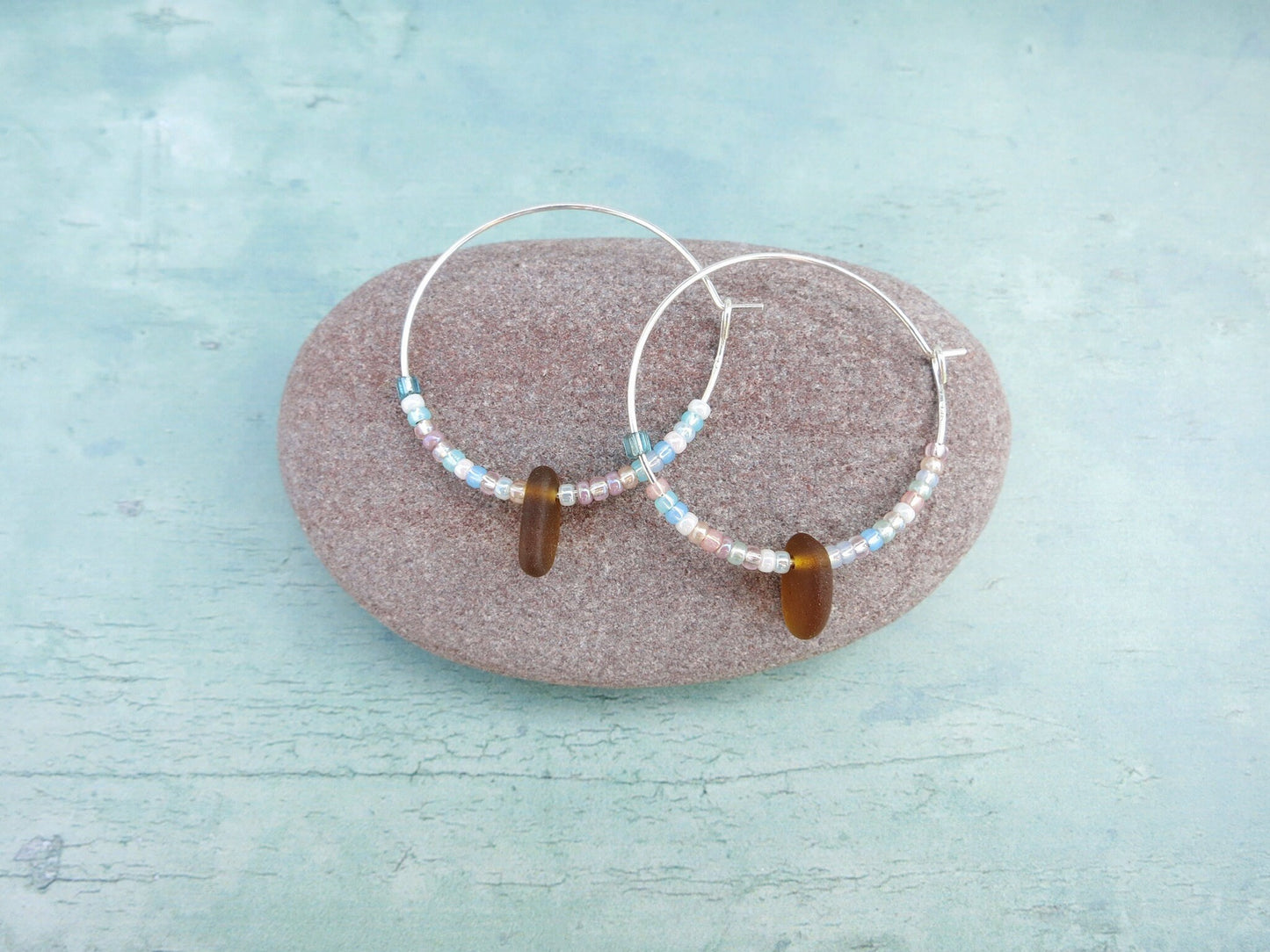 Silver hoop earrings with pastel seed beads and burnt orange sea glass laid on a light blue background