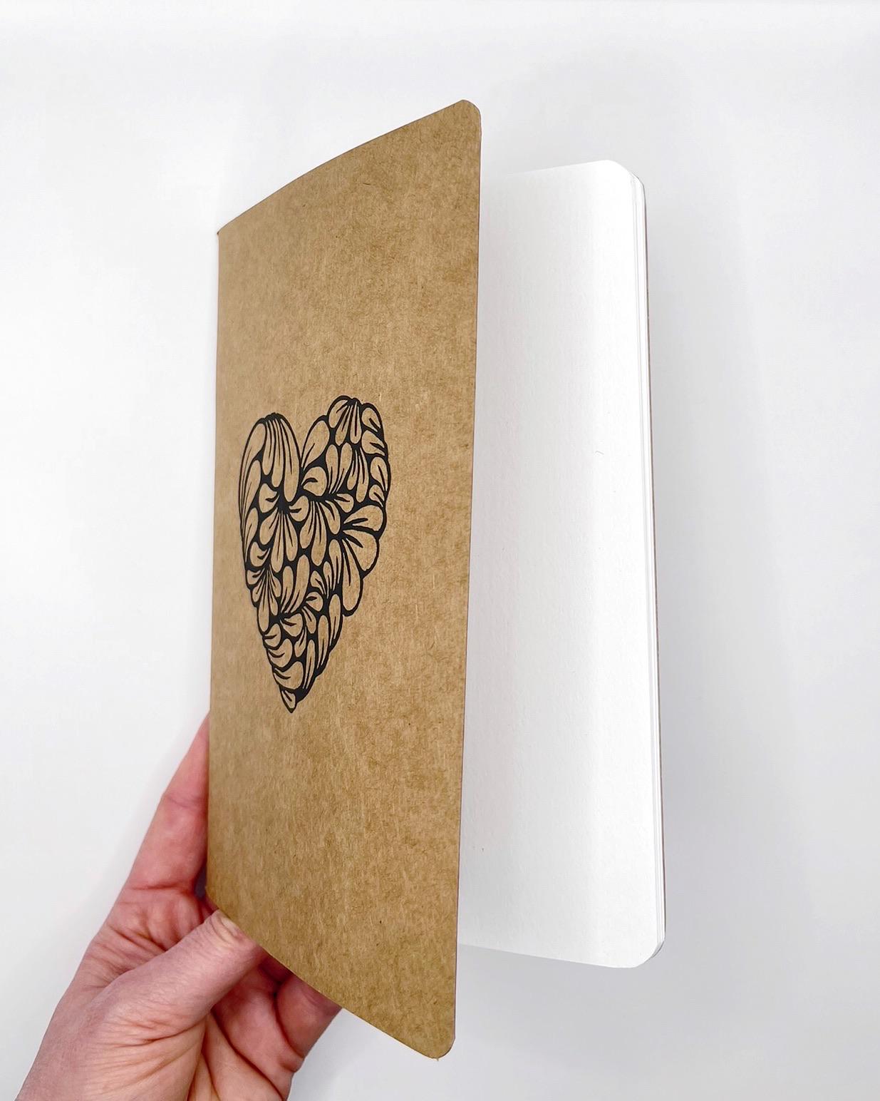 Kraft paper notebook with a heart design held by a hand on a white background showing the plain white pages