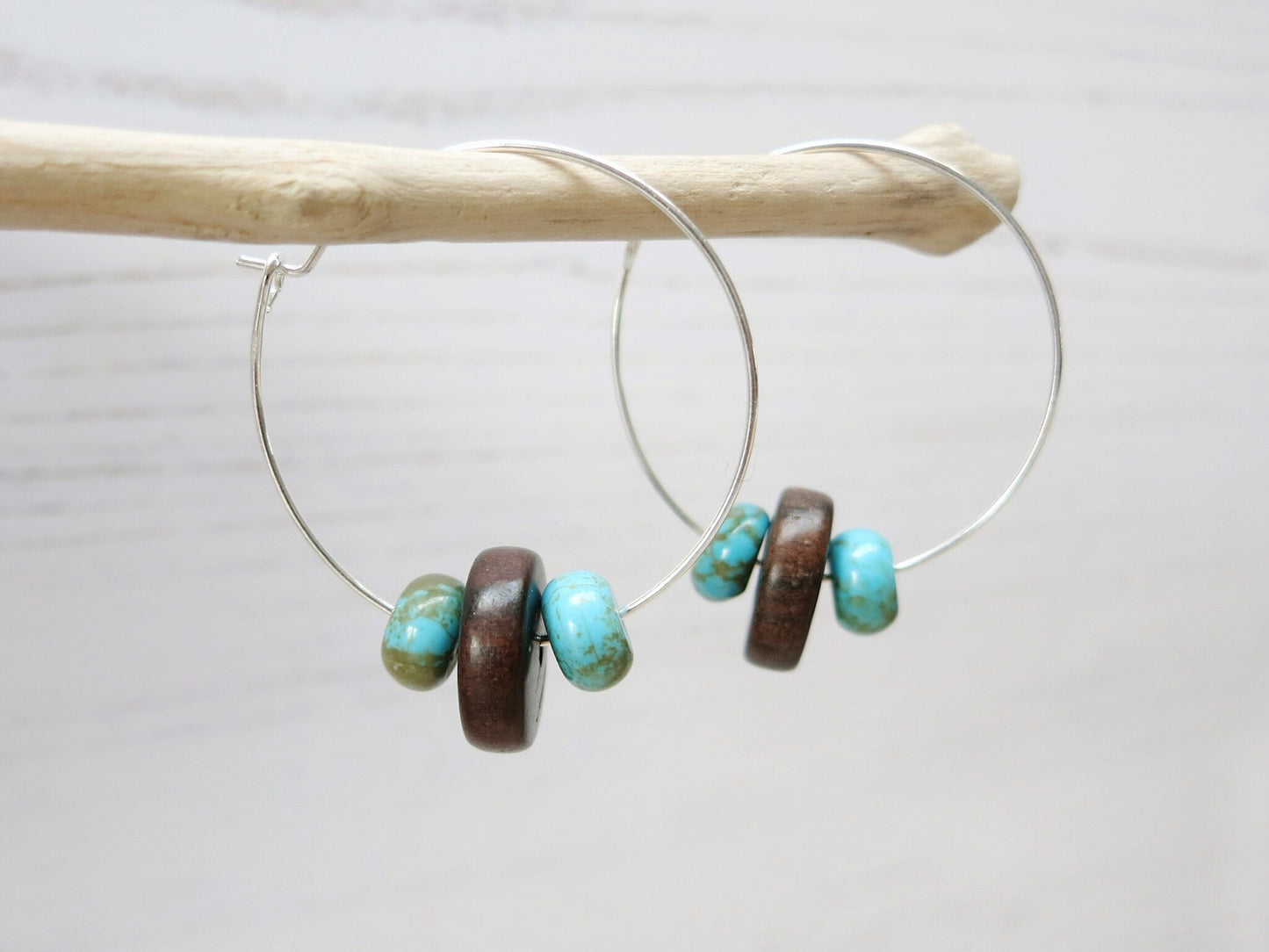 Close up of silver hoop earrings with turquoise and wooden beads hanging on driftwood against a light background
