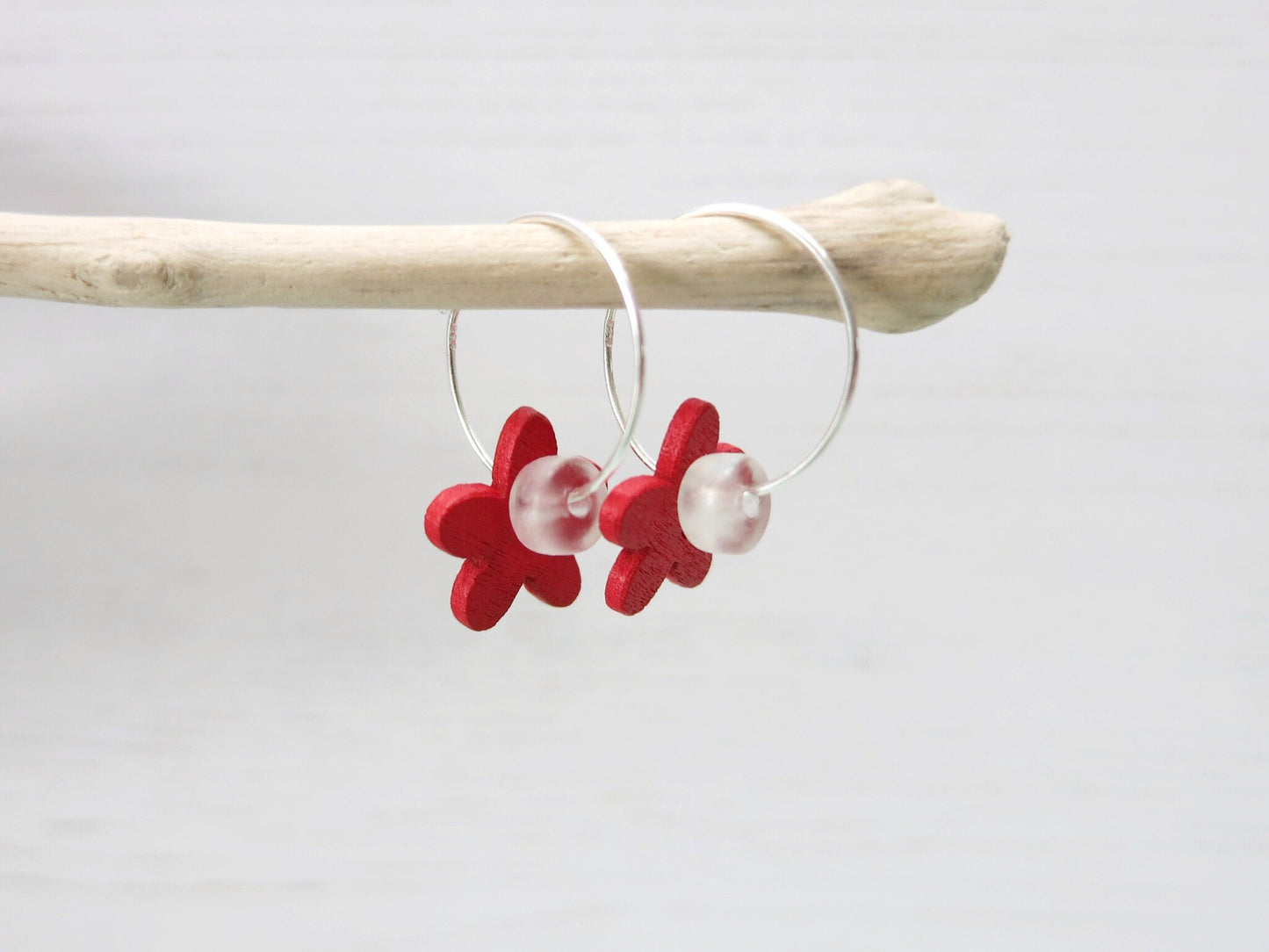 Side view of silver hoop earrings with red wooden flowers and a glass bead hanging on driftwood against a white background