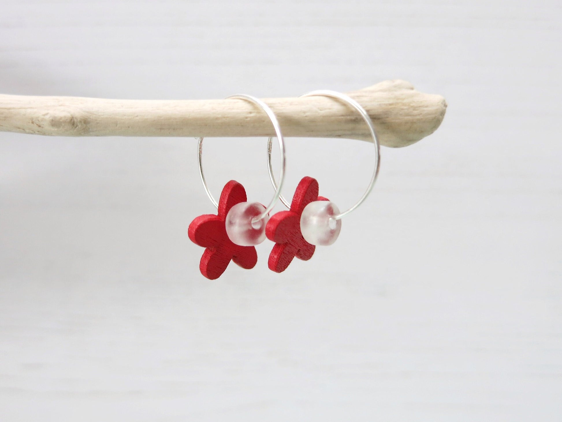 Side view of silver hoop earrings with red wooden flowers and a glass bead hanging on driftwood against a white background