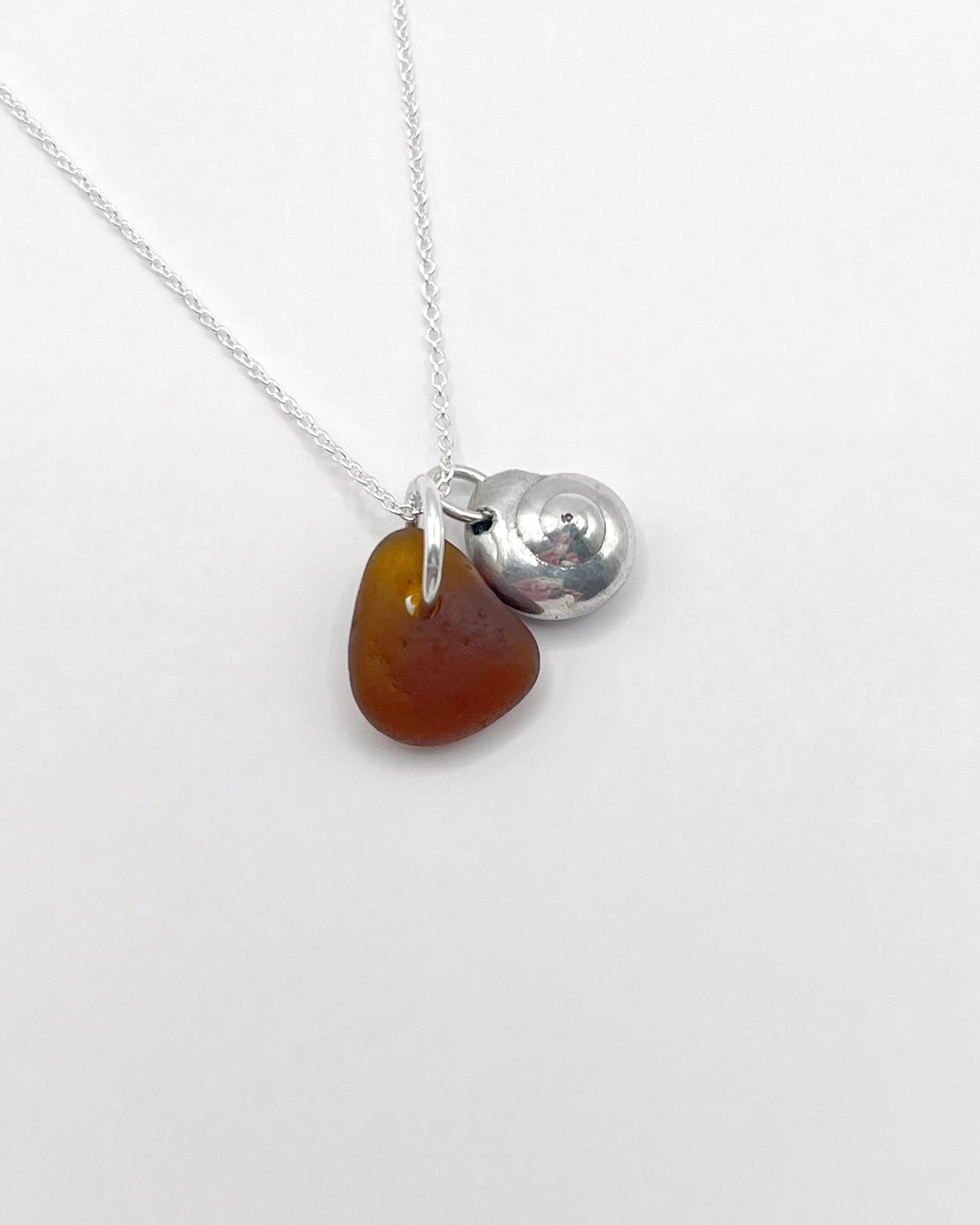 Necklace with burnt orange sea glass and a silver shell pendant, laid on a white surface