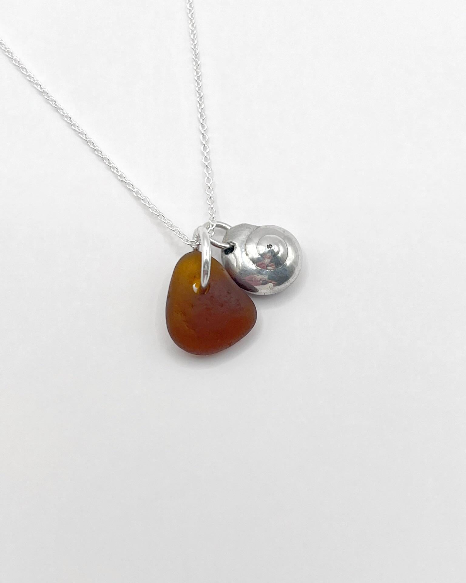 Necklace with burnt orange sea glass and a silver shell pendant, laid on a white surface
