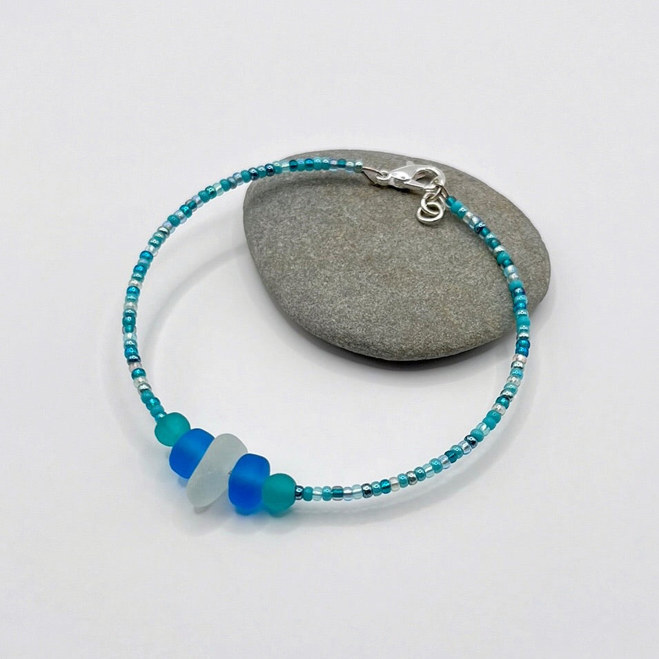 Bracelet with white sea glass and blue and turquoise beads laid on a grey pebble against a white background
