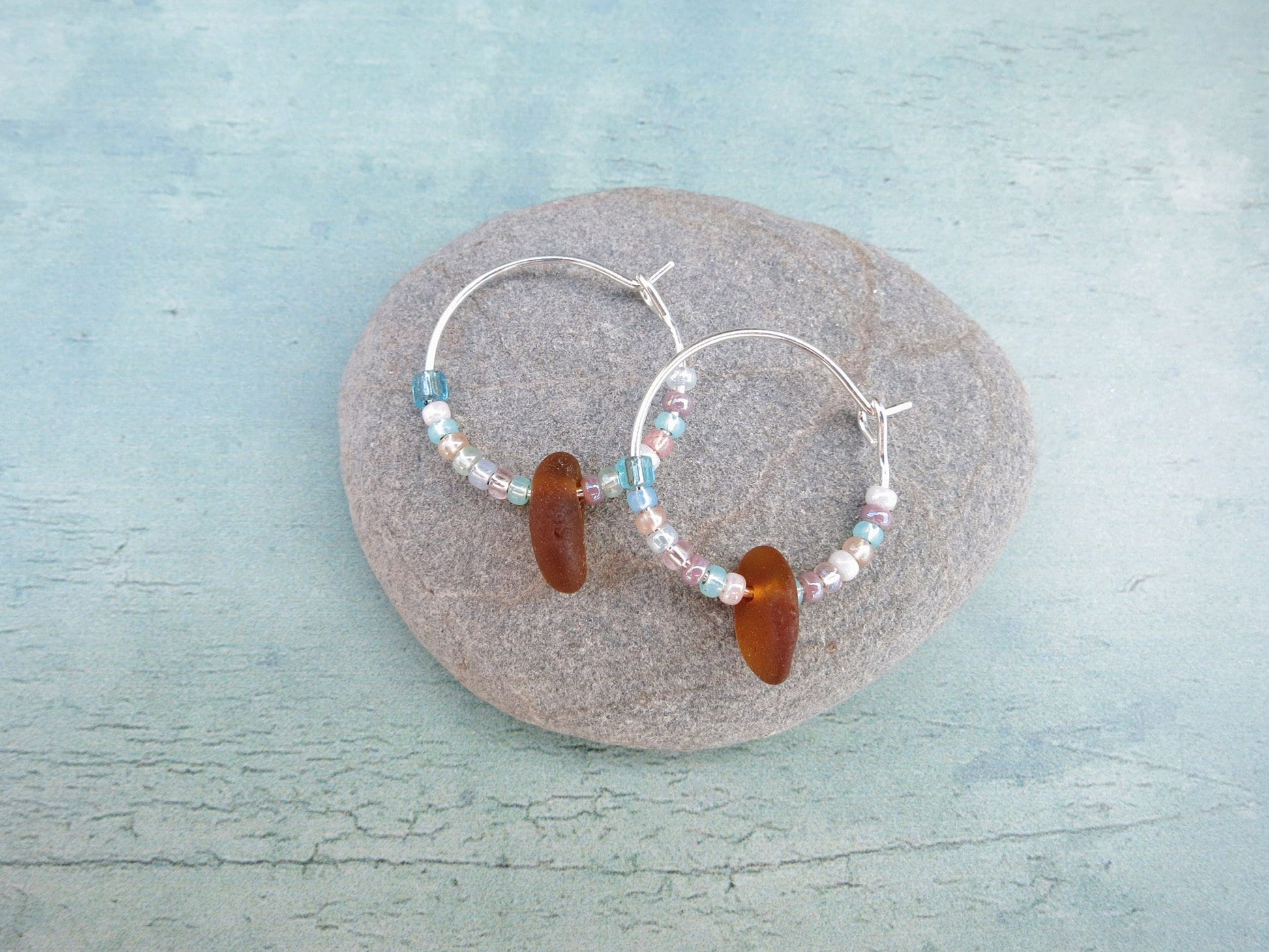 Silver hoop earrings with pastel seed beads and burnt orange sea glass, laid on a grey pebble against a light blue background