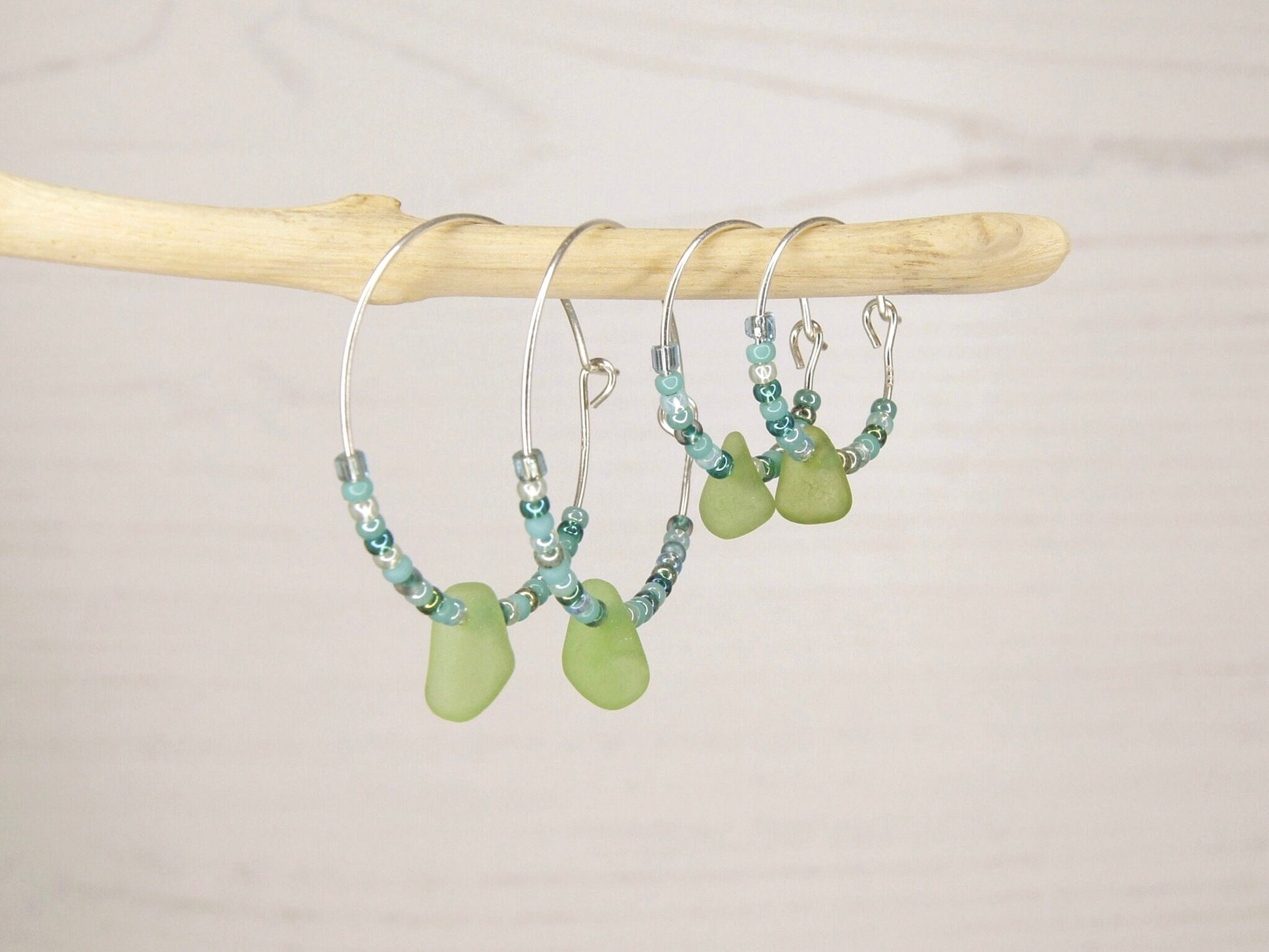 Hoop earrings with green sea glass and turquoise seed beads hanging on driftwood  against a pale background