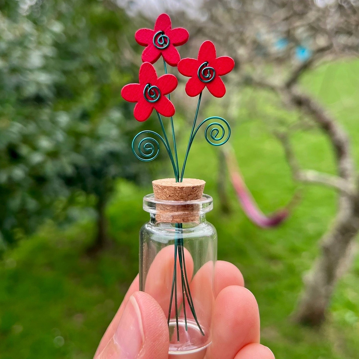 Little glass bottle with three red wooden flowers held in a hand against a garden background