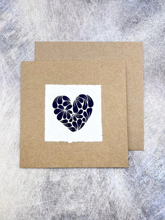 Kraft card with original lino printed blue heart design on white paper with brown envelope laid on a textured grey background