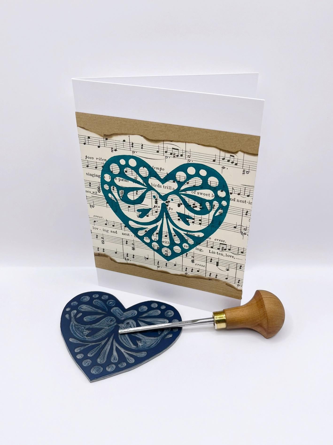 Card with a green lino printed heart design on sheet music. Photo includes lino and cutting tool laid on a white background.