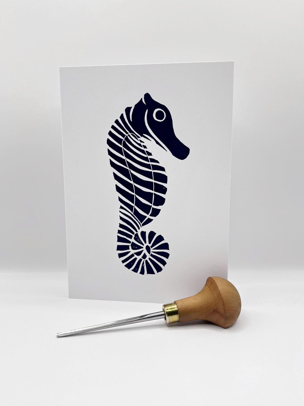 Original lino print of a navy blue seahorse design on a white card, stood against a white background. Photo includes lino cutting tool.