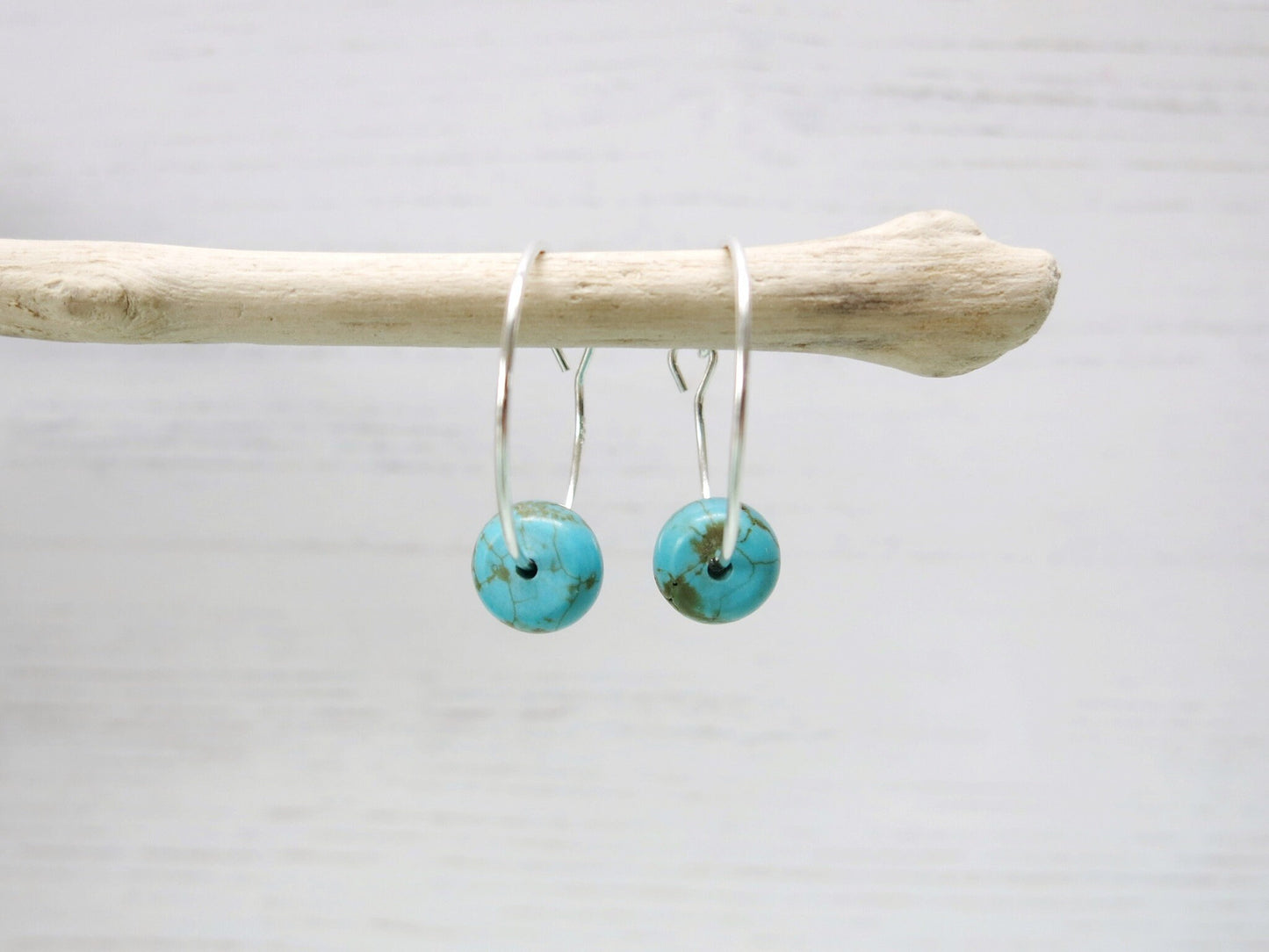 Close up of silver hoop earrings with turquoise beads hanging on driftwood against a pale background