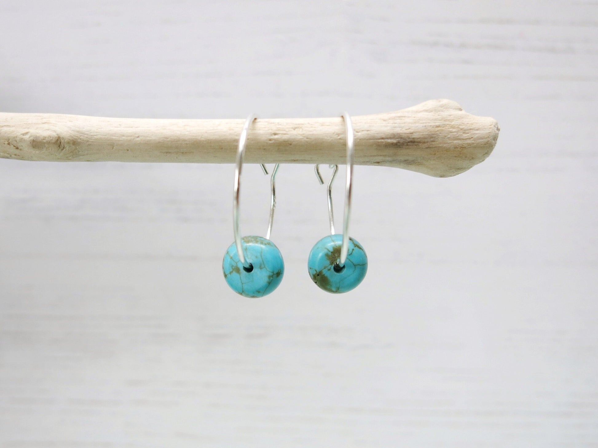 Close up of silver hoop earrings with turquoise beads hanging on driftwood against a pale background