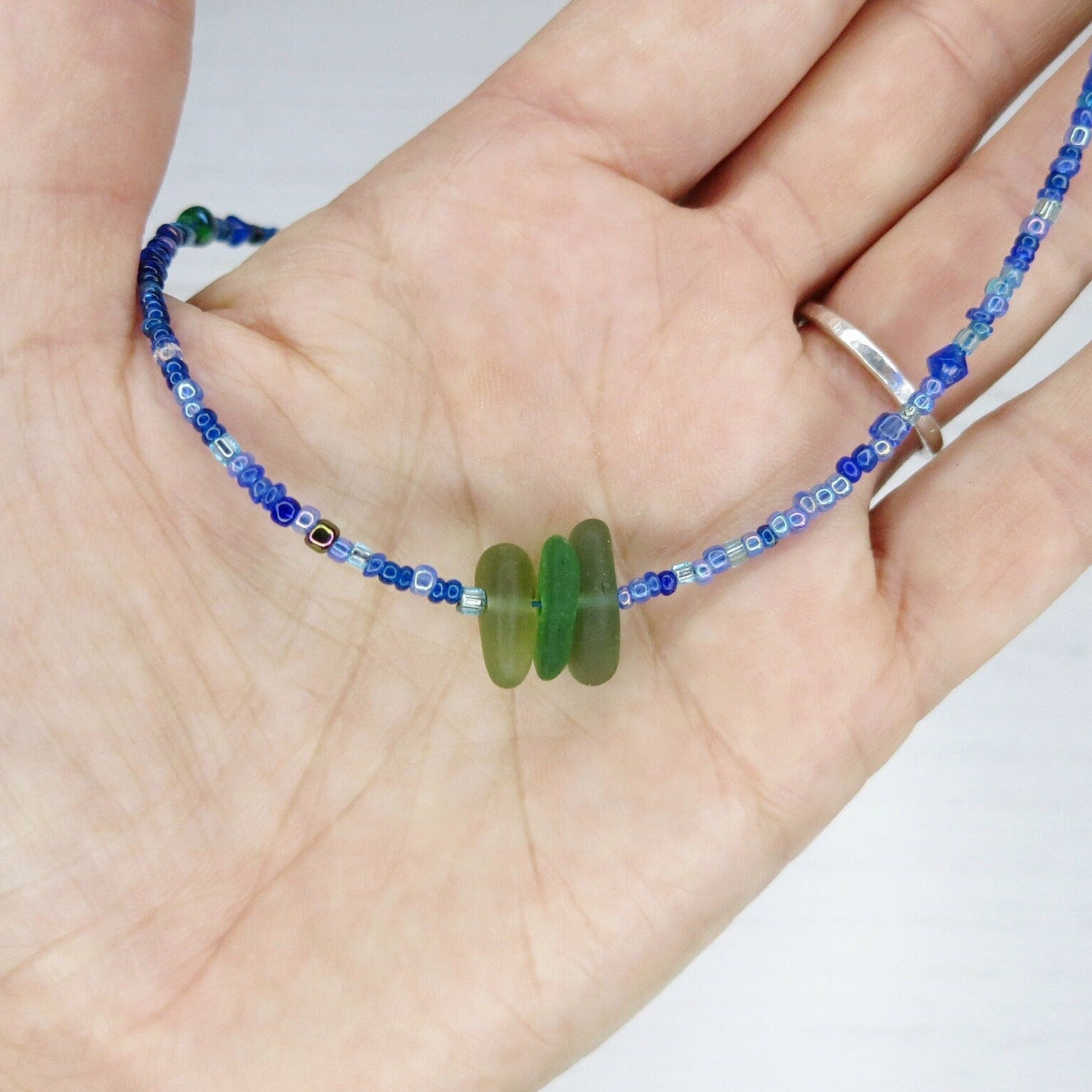 Hand holding a blue seed bead necklace with green sea glass against a plain background