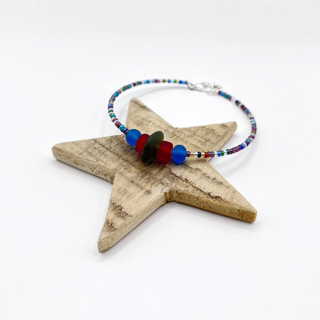 A rainbow coloured beaded bracelet with red and blue frosted glass beads and a piece of olive green coloured Cornish sea glass, finished with a silver plated lobster clasp and laid on a wooden star.