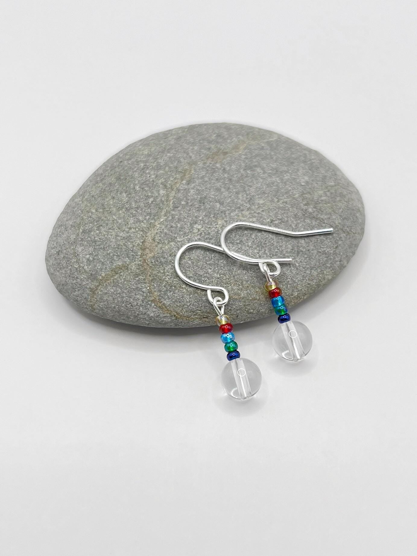 Silver earrings with clear crystal and rainbow seed beads laid on a grey pebble