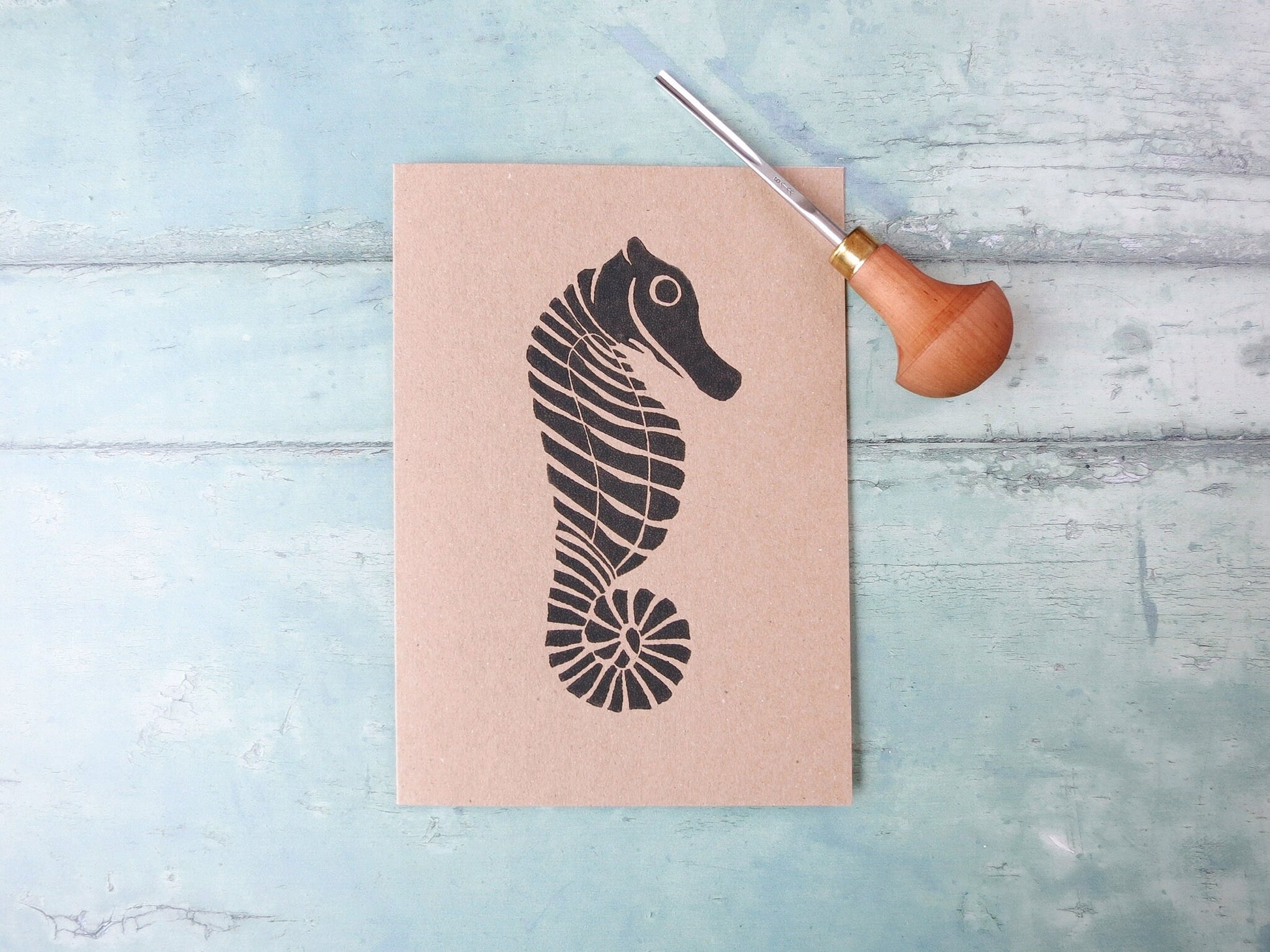 Kraft paper card with an original seahorse lino print laid on a textured blue surface. Photo includes lino cutting tool