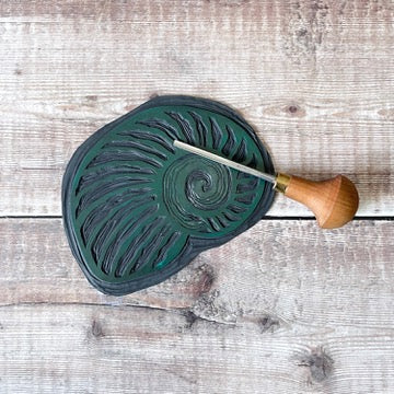 The lino used for the seashell printed wash bag. Photo includes the lino cutting tool used and is laid on a wooden background.